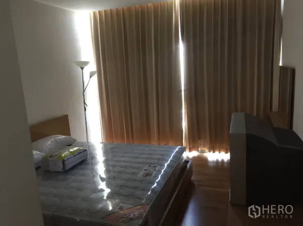 Condominium for rent in Sathon, Bangkok - Bedroom with blackout curtains, new mattress on the bed, floor lamp, and TV console on timber flooring.