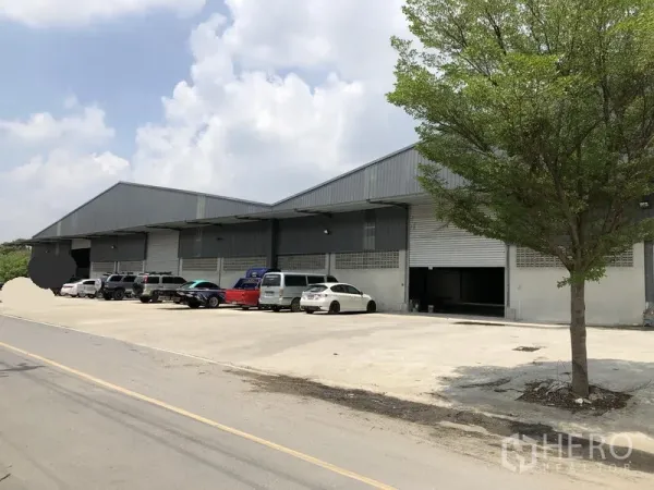 คลังสินค้า ให้เช่าในBang Kruai, Nonthaburi - ภาพจากถนนของอาคารโกดังขนาดใหญ่พร้อมลานคอนกรีตและต้นไม้