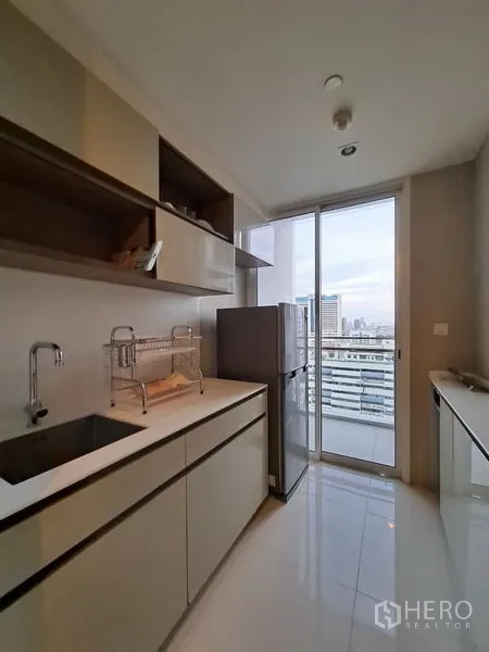公寓 出租在Bang Rak, Bangkok - 廊形厨房，简洁橱柜与水槽，落地推拉门通往阳台眺望天际线。