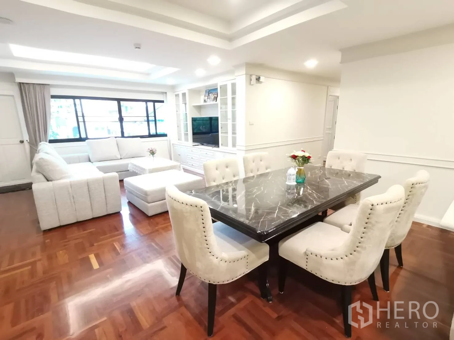 Condominium for rent in Watthana, Bangkok - Dining area with marble-top table and upholstered chairs beside a bright lounge with sectional sofa and large window.