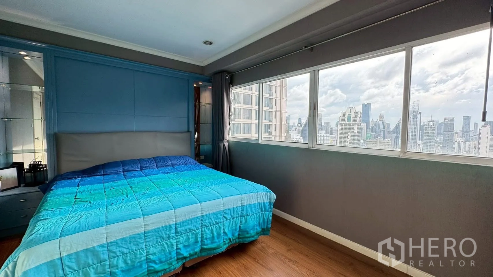 Condominium for rent or sale in Watthana, Bangkok - Master bedroom with blue headboard and wide windows facing the Bangkok city skyline.