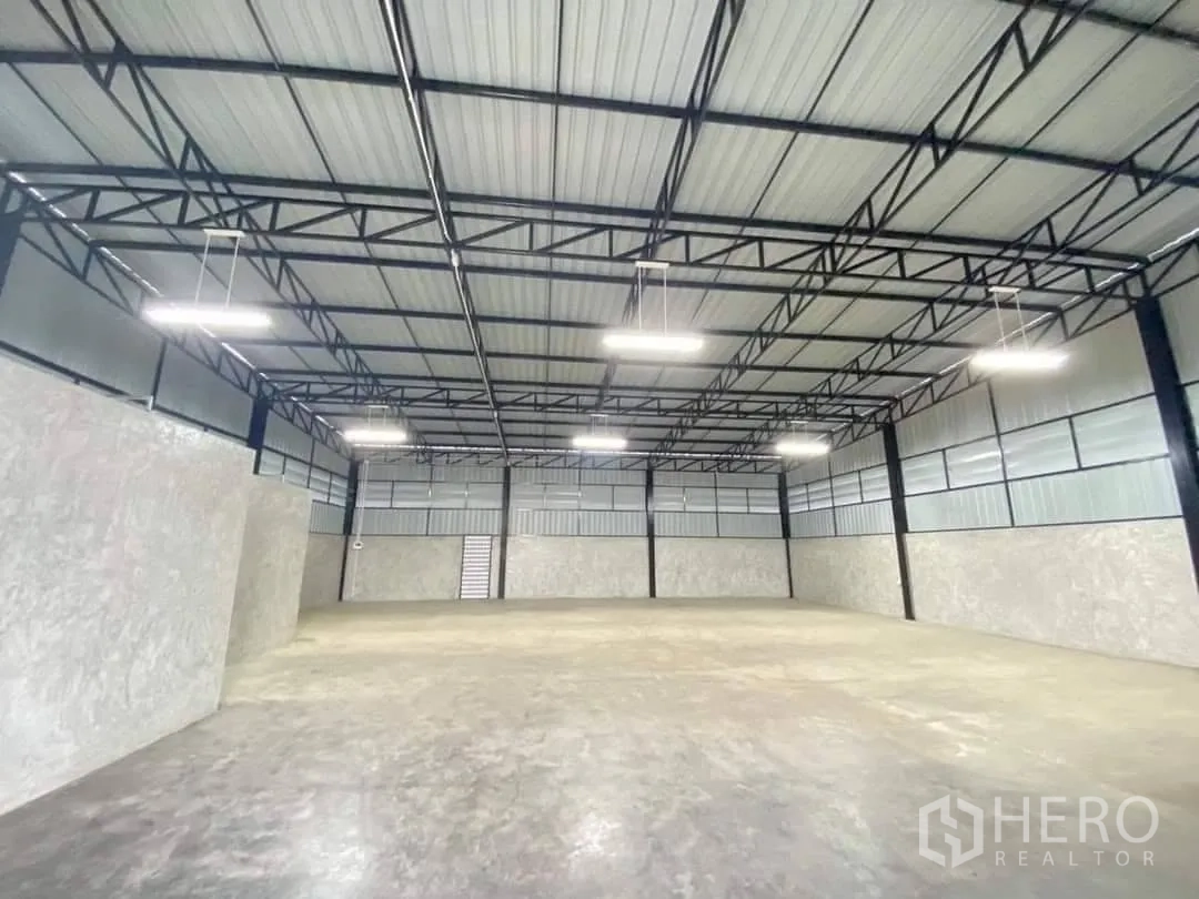 Warehouse for rent in Bang Bua Thong, Nonthaburi - Open-plan warehouse interior with steel roof trusses, hanging lights, and a smooth concrete floor.