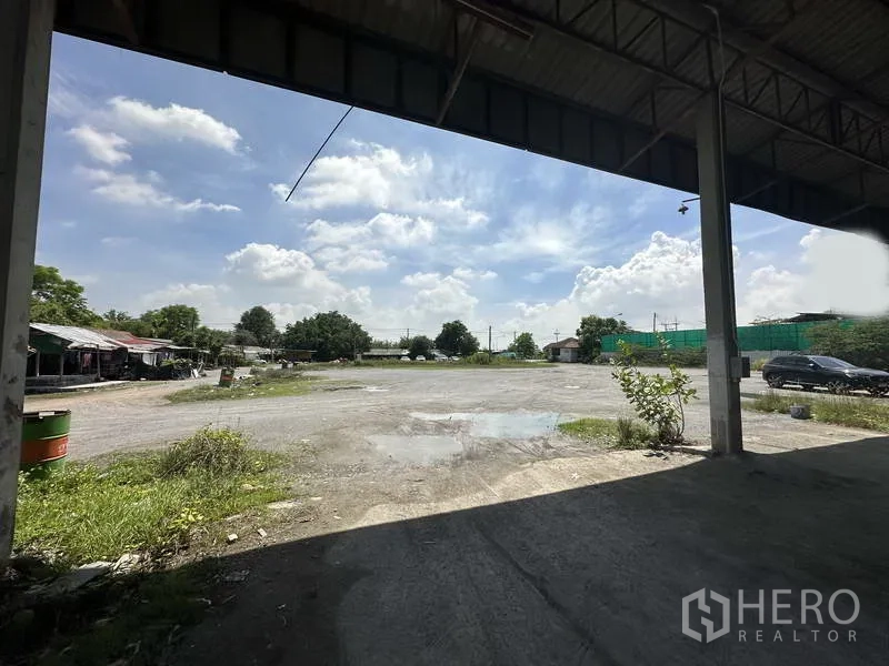 工厂 出租在Phra Nakhon Si Ayutthaya - 在屋檐下眺望开阔的碎石场地，朝向场地入口与树木。