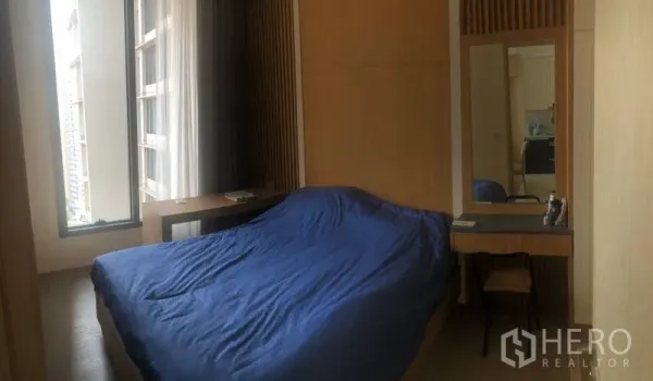 Condominium for rent or sale in Watthana, Bangkok - Cozy second bedroom with desk, mirror and floor-to-ceiling window overlooking city buildings.