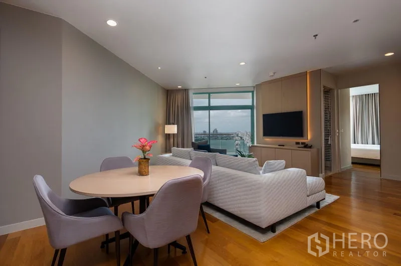 Condominium for rent in Bang Kho Laem, Bangkok - Open-plan living and dining area with round table, L-shaped sofa and river view through floor-to-ceiling windows in a modern Bangkok condo.