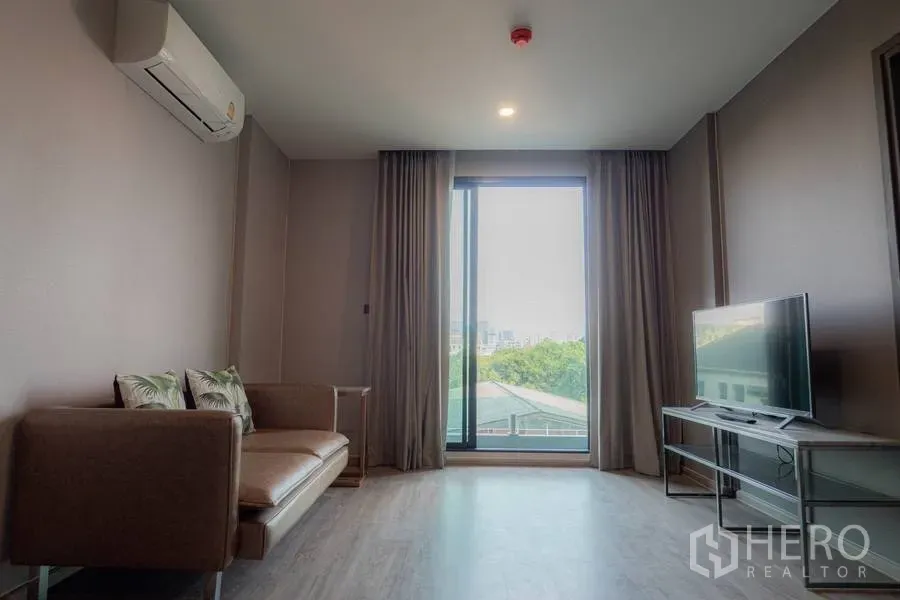 Condominium for rent or sale in Watthana, Bangkok - Bright living room with sofa, TV console, and large sliding window overlooking greenery and skyline.