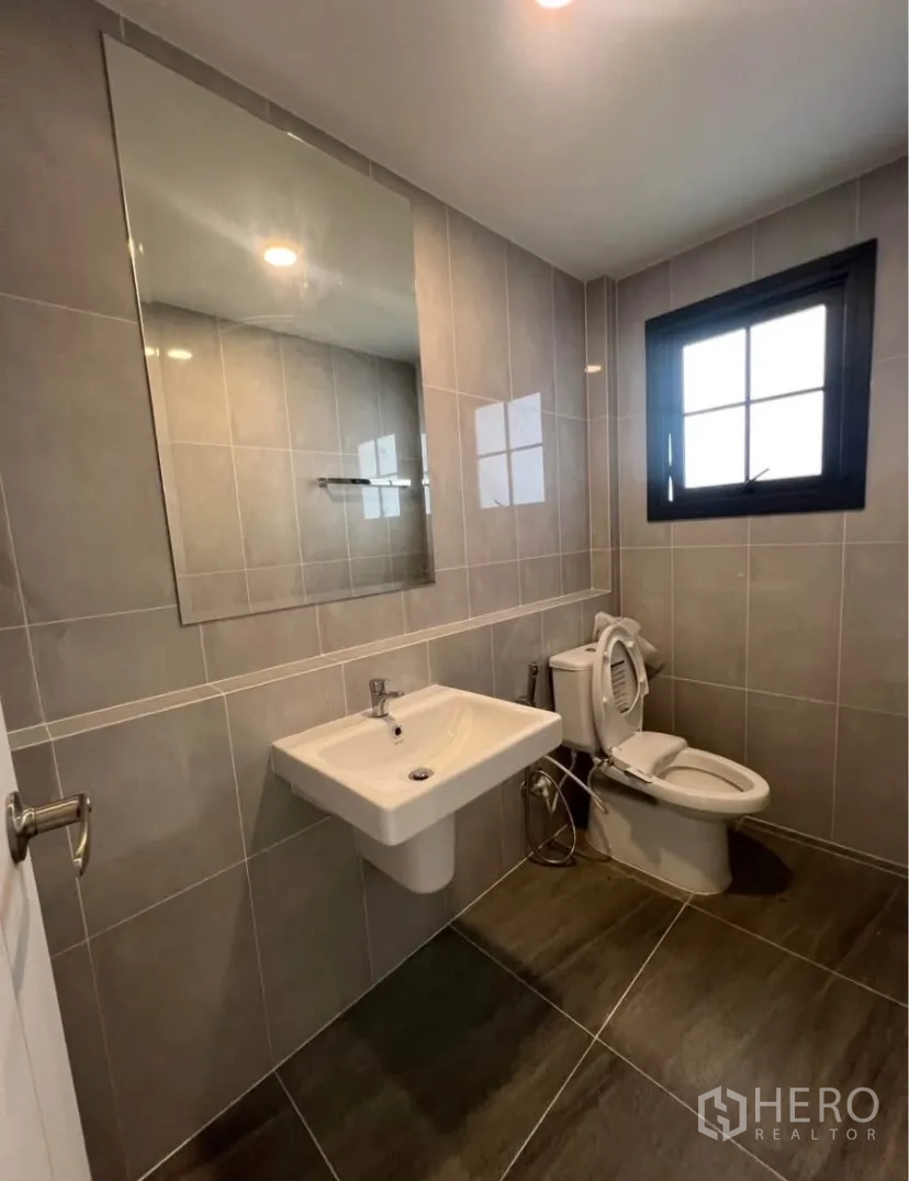 Detached House for rent in Bang Phli, Samut Prakan - Tiled bathroom featuring a large mirror, pedestal sink, toilet and a window for natural light.