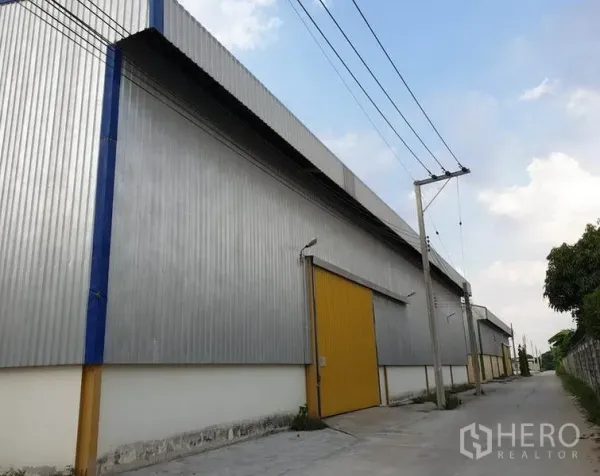 货栈 出租在Lat Krabang, Bangkok - 金属板外墙仓库侧立面，黄色卷帘门，沿安静小路与电线杆。