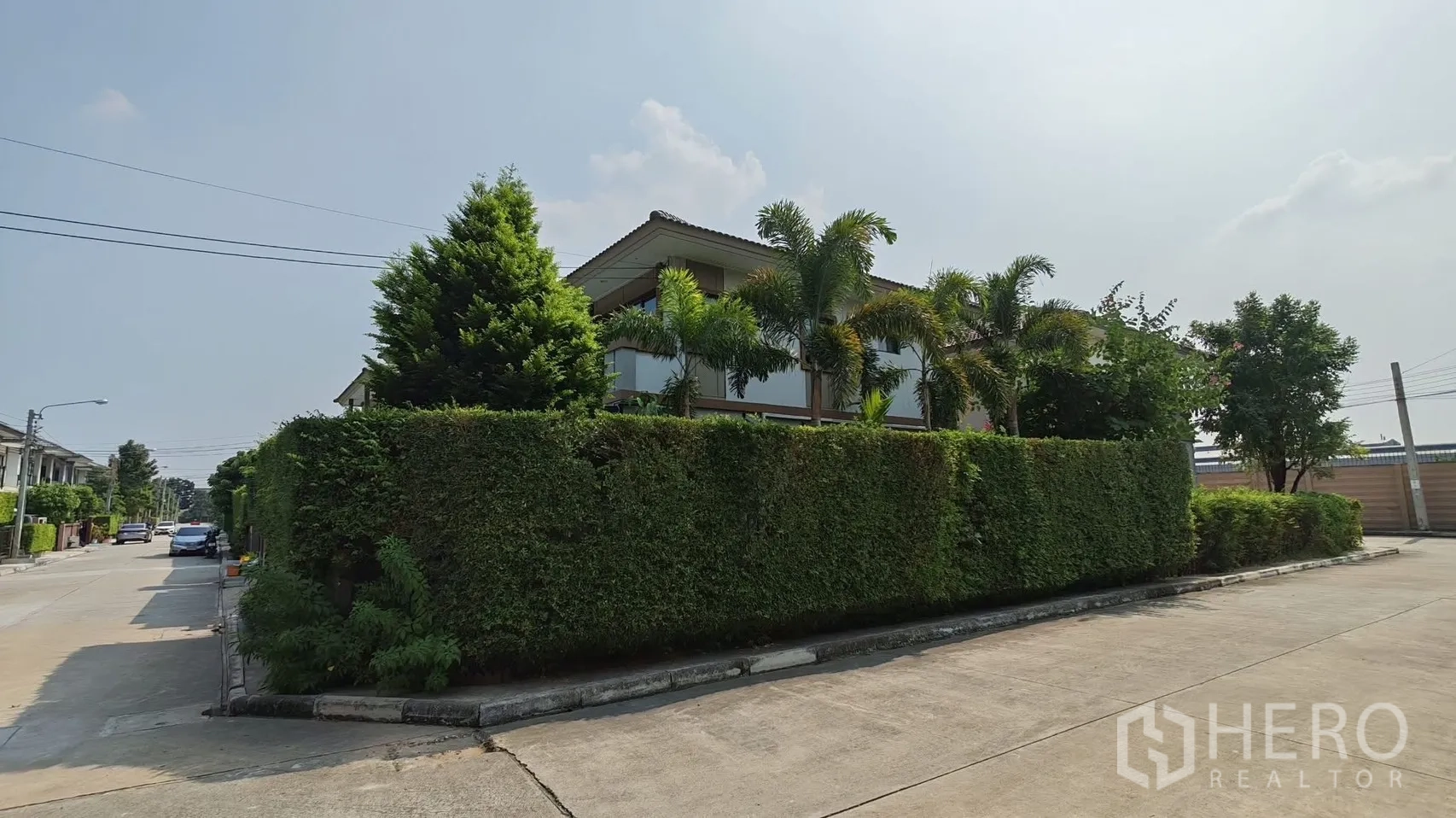 独栋房屋 for rent or sale in Bang Phli, Samut Prakan - 被高大绿篱与棕榈环绕的两层转角独栋外观，临安静社区道路。