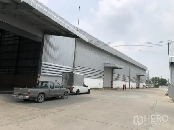 Warehouse for rent in Pathum Thani - Large corrugated-steel warehouse exterior with multiple loading bays and parking on a wide concrete driveway in Pathum Thani.