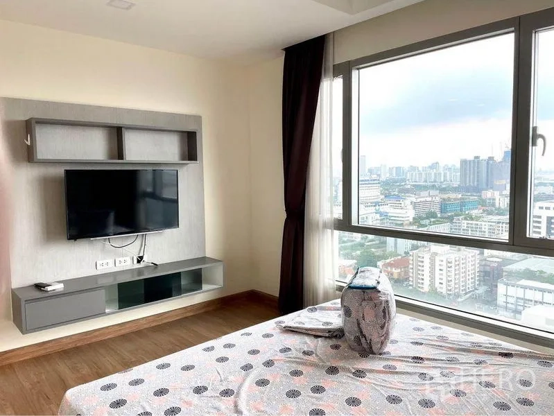 Condominium for rent in Huai Khwang, Bangkok - Bedroom with TV wall unit and panoramic Bangkok skyline through wide windows.
