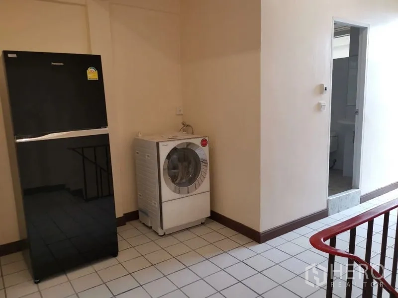 Townhouse for rent in Watthana, Bangkok - Utility area with black fridge, front-load washing machine, and doorway to a bathroom.