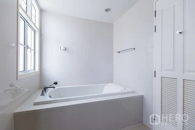 Townhouse for rent in Watthana, Bangkok - Bathroom with built-in bathtub next to a tall window and neutral tiles.