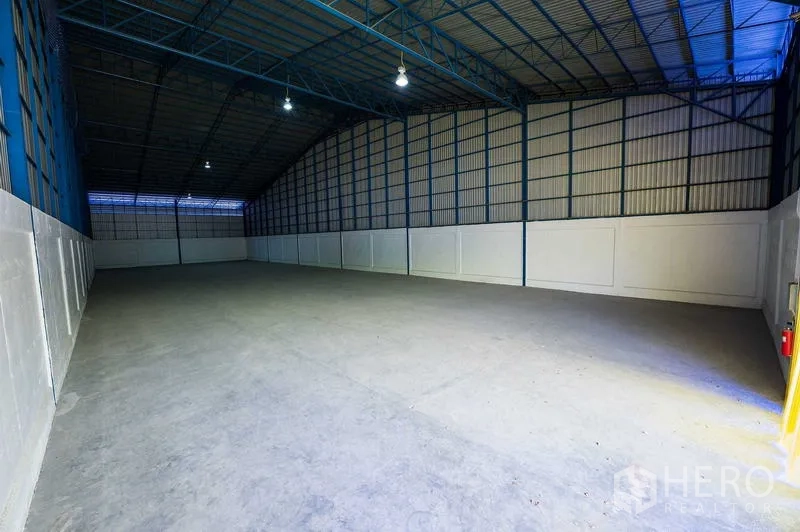Warehouse for rent in Phra Phutthabat, Saraburi - Clean empty storage bay with high roof, white lower wall panels, and a fire extinguisher by the door.