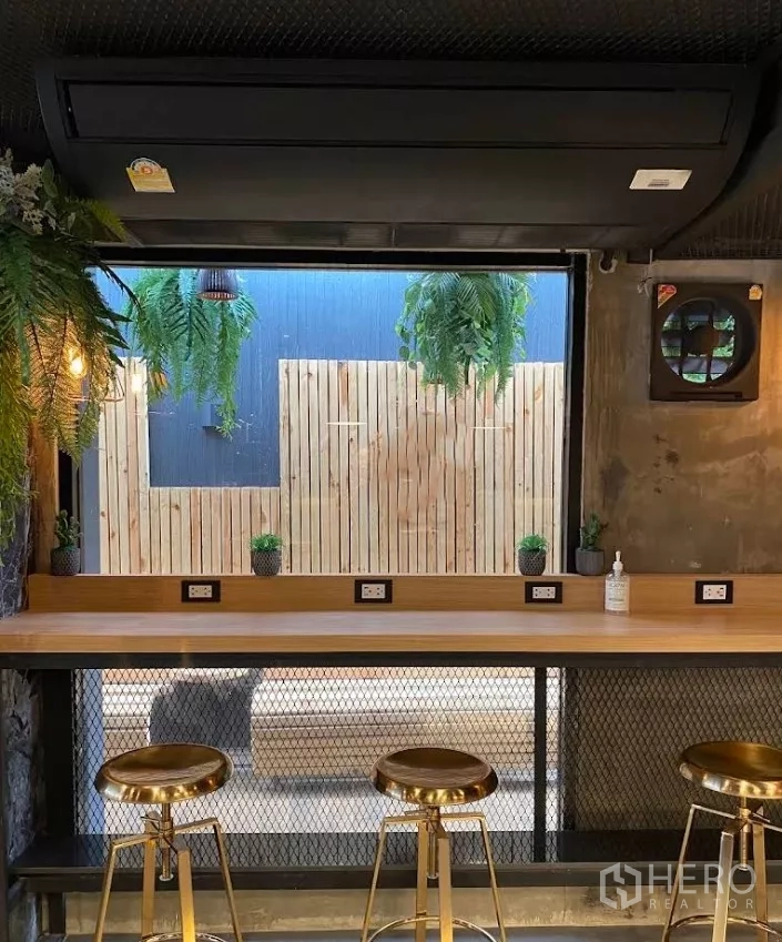 พื้นที่ขายปลีก ให้เช่าในKhlong Toei, Bangkok - พื้นที่ค้าปลีกภายในพร้อมเคาน์เตอร์ไม้ยาว ช่องเสียบปลั๊ก และสตูลสีทอง มองออกสู่ลานต้นไม้