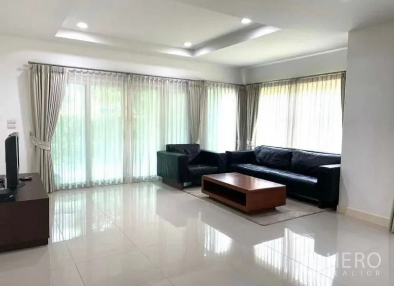 บ้าน ให้เช่าหรือขายในLat Krabang, Bangkok - ห้องนั่งเล่นสว่างด้วยหน้าต่างมุมกว้าง ผ้าม่านโปร่ง โซฟาหนังสีดำและโต๊ะไม้