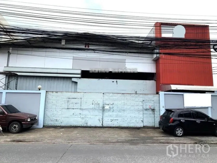 Warehouse for rent in Suan Luang, Bangkok - Street-front warehouse exterior in Suan Luang, Bangkok with high boundary wall, wide metal gate and overhead utility lines.