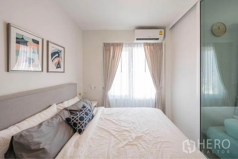 Condominium for sale in Huai Khwang, Bangkok - Cozy bedroom with large bed, air conditioning, and a sheer-curtained window letting in soft daylight.