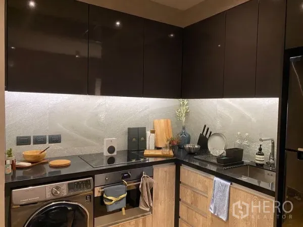 Condominium for rent or sale in Watthana, Bangkok - Compact L-shaped kitchen with black stone countertop, induction hob, built-in oven, washer, and glossy cabinets.