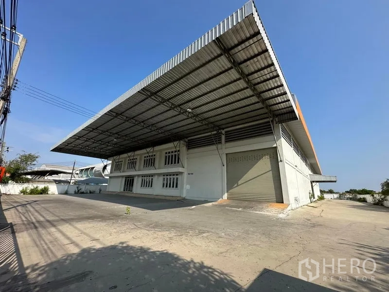 Factory for rent in Samut Prakan - Large factory exterior in Bang Bo with wide steel canopy, roller shutter door, and spacious paved yard for truck loading.