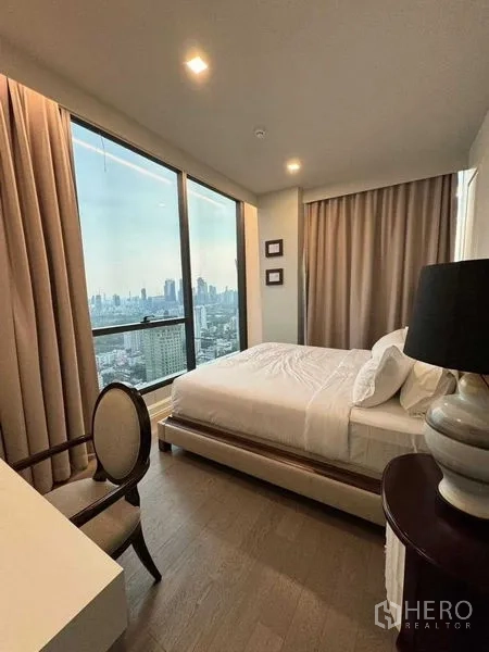 Condominium for rent in Watthana, Bangkok - Master bedroom with corner windows, sweeping city panorama, and a desk beside a bedside lamp.