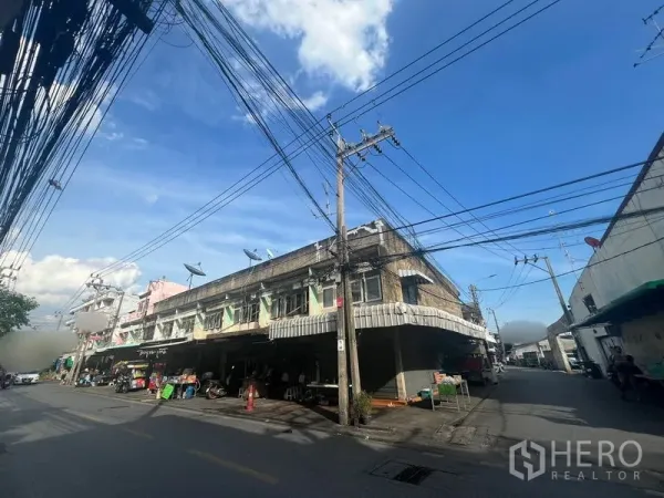 ที่ดิน ให้เช่าหรือขายในPhra Khanong, Bangkok - ภาพกว้างของตึกแถวหัวมุม ติดถนนสองด้าน มีสายไฟเหนือศีรษะในบรรยากาศตลาดชุมชน.