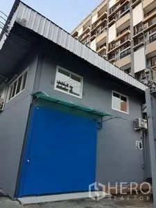Warehouse for rent in Chatuchak, Bangkok - Compact gray warehouse corner featuring a blue entry door, awning and corrugated roof in an urban Chatuchak setting.