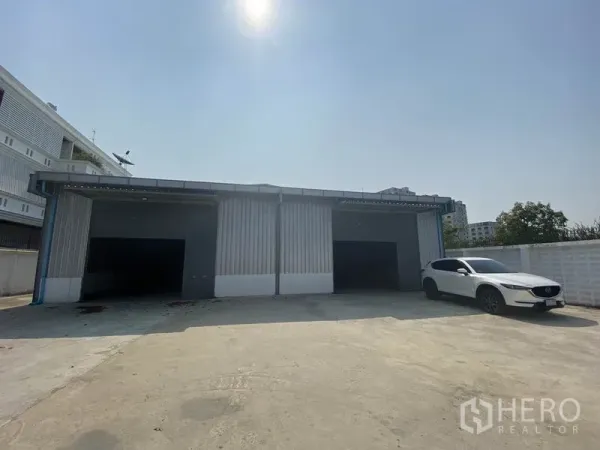 货栈 出租在Prawet, Bangkok - 金属外墙仓库外观，三扇卷帘门与宽阔混凝土院场，旁停一辆白色SUV。