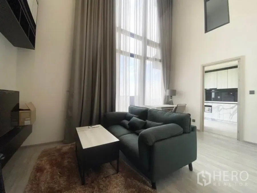 Condominium for rent in Phra Khanong, Bangkok - Double-height living room with dark sofa, tall curtains and doorway to a bright kitchen in a modern Bangkok condo.