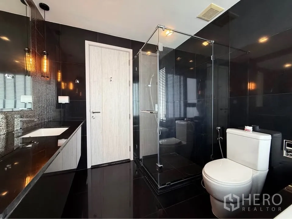 Condominium for rent in Khlong Toei, Bangkok - Stylish black-tiled bathroom with glass shower, toilet and long vanity counter.