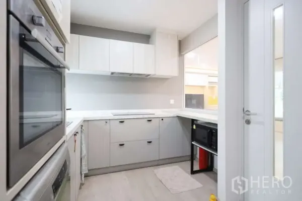 Detached House for rent or sale in Prawet, Bangkok - Light-toned L-shaped kitchen with built-in appliances, ample cabinets and a window.