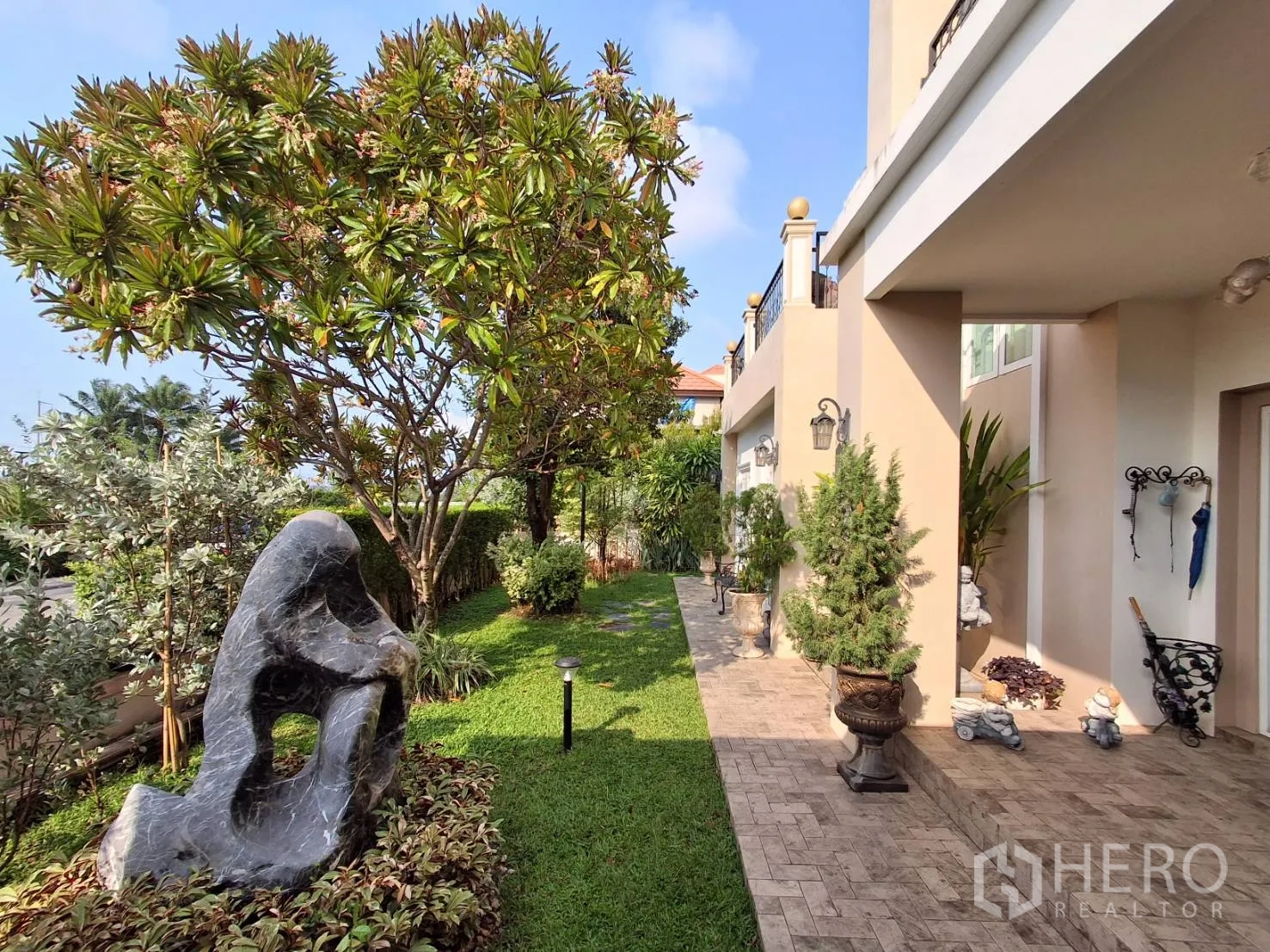Detached House for rent or sale in Ban Chang, Rayong - Side garden with paved walkway, trees, shrubs and decorative stone sculpture.