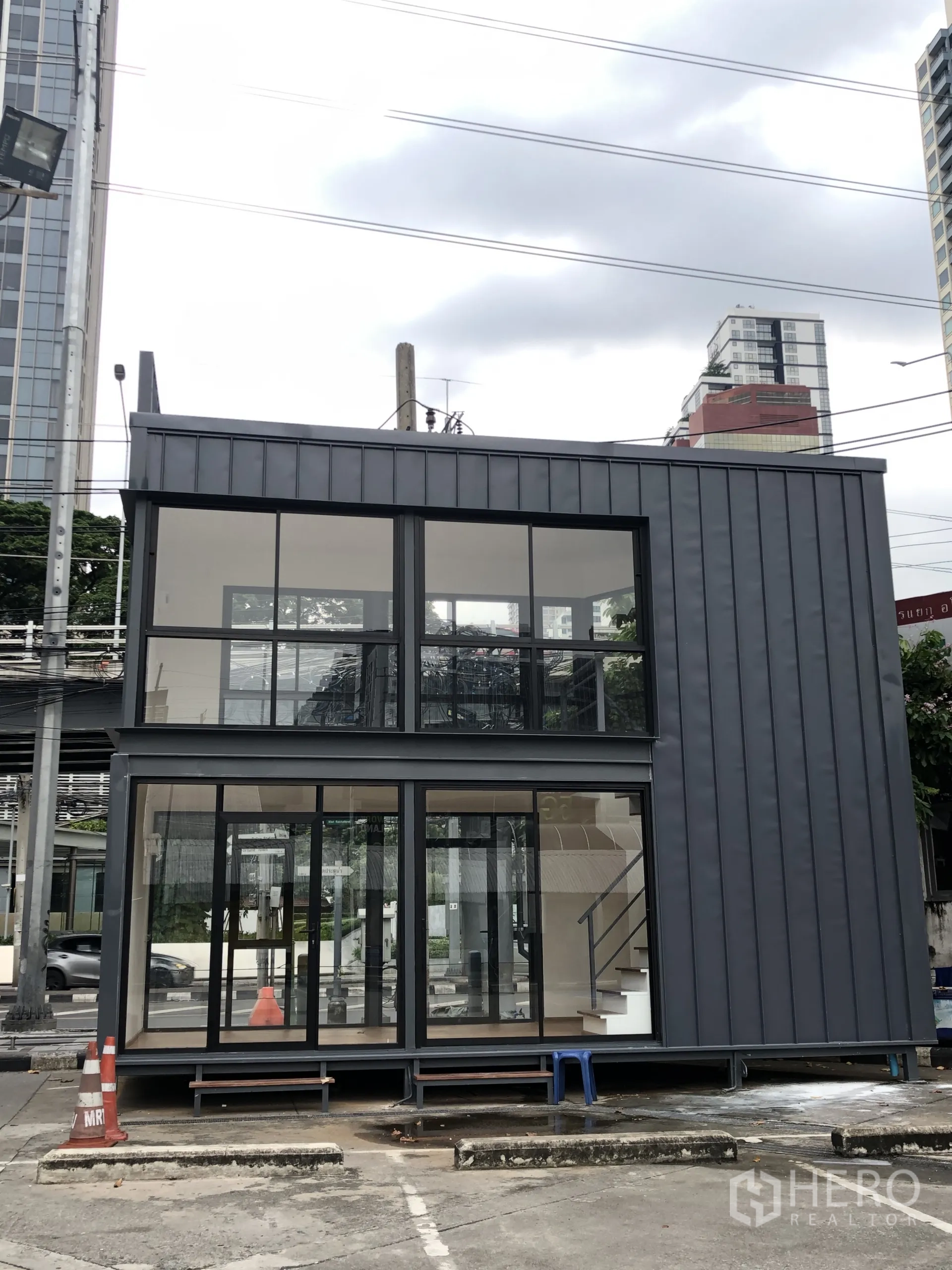 零售空间 出租在Ratchathewi, Bangkok - 双层零售铺面，落地玻璃立面与深色金属外墙，室内楼梯，临街入口，位于曼谷城市街景中。