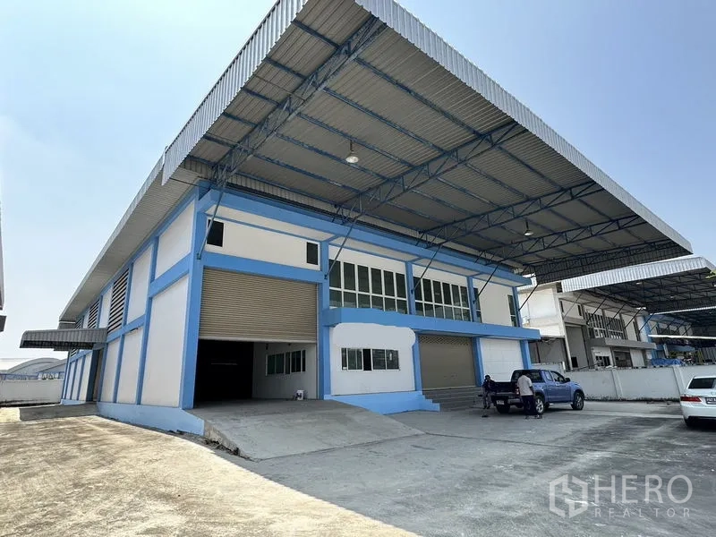 Factory for rent in Bang Bo, Samut Prakan - Blue-and-white factory exterior with wide metal canopy, loading ramp and roller shutters in an industrial compound.