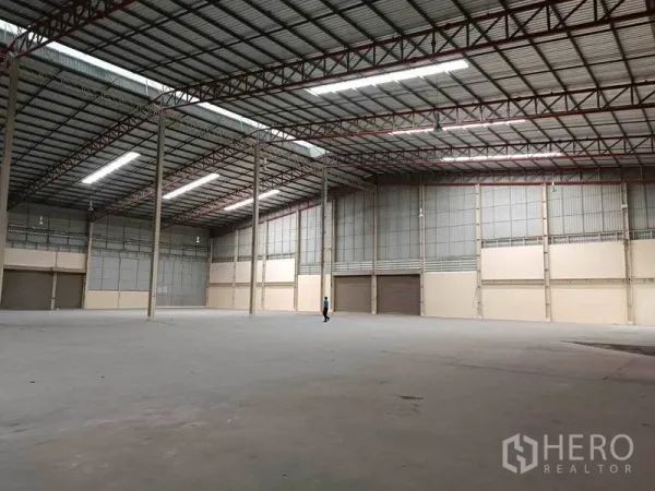 工厂 出租在Samut Prakan - 邦普里大型厂库内部，高挑钢架屋顶、明亮灯带、混凝土地面与多处卷帘门装卸口。