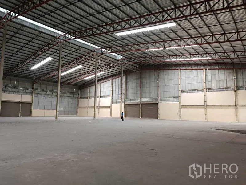工厂 出租在Samut Prakan - 邦普里大型厂库内部，高挑钢架屋顶、明亮灯带、混凝土地面与多处卷帘门装卸口。