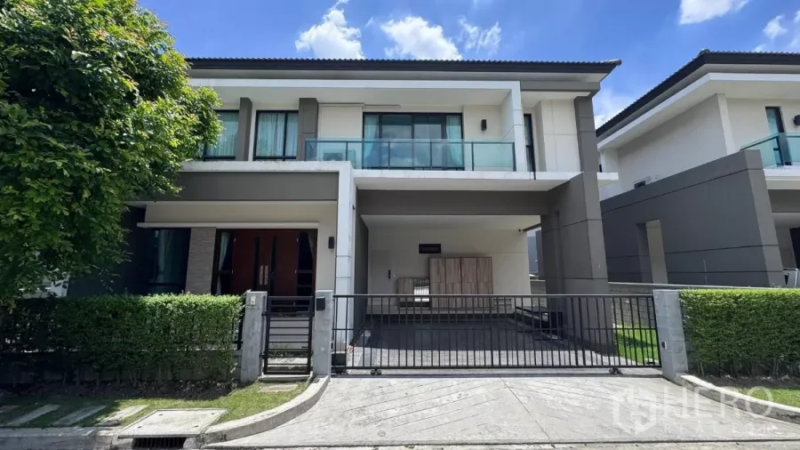 บ้าน ให้เช่าหรือขายในSaphan Sung, Bangkok - บ้านเดี่ยวสองชั้นสไตล์โมเดิร์นในเขตสะพานสูง กรุงเทพฯ มีรั้วหน้าบ้าน ที่จอดรถ และระเบียงกระจก รายล้อมด้วยแนวพุ่มไม้ท่ามกลางท้องฟ้าโปร่งใส.