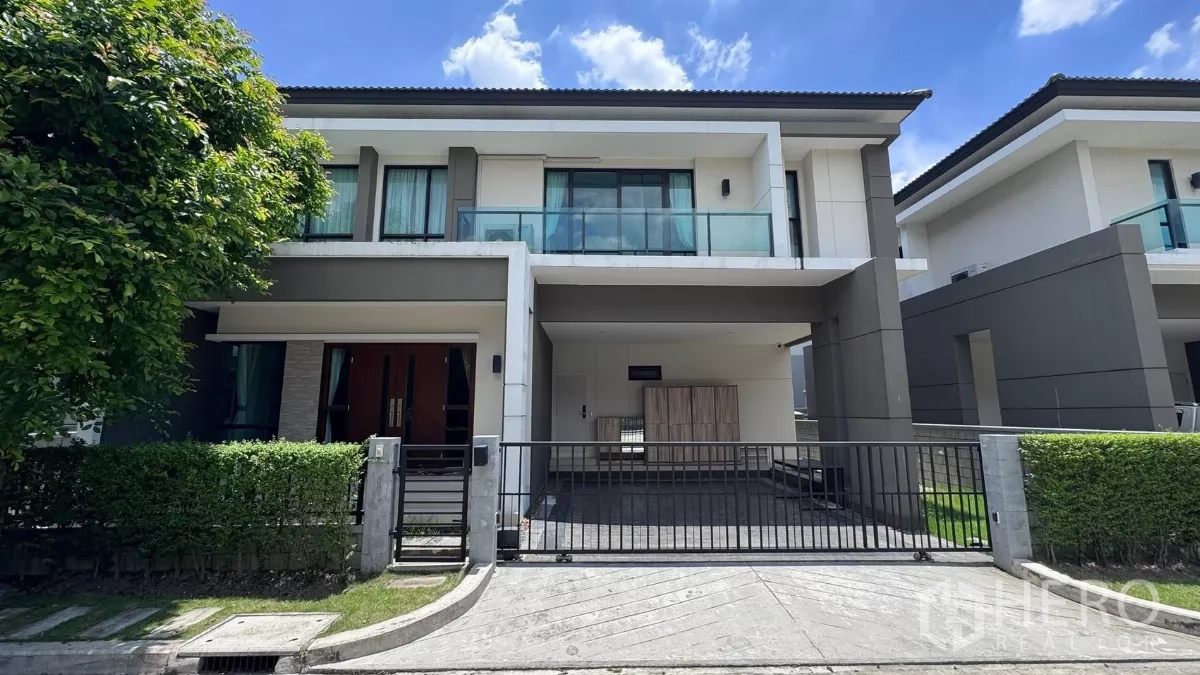 บ้าน ให้เช่าหรือขายในSaphan Sung, Bangkok - บ้านเดี่ยวสองชั้นสไตล์โมเดิร์นในเขตสะพานสูง กรุงเทพฯ มีรั้วหน้าบ้าน ที่จอดรถ และระเบียงกระจก รายล้อมด้วยแนวพุ่มไม้ท่ามกลางท้องฟ้าโปร่งใส.