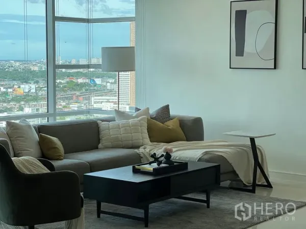Condominium for rent in Watthana, Bangkok - Bright living area with grey sectional sofa and floor‑to‑ceiling windows overlooking Bangkok.