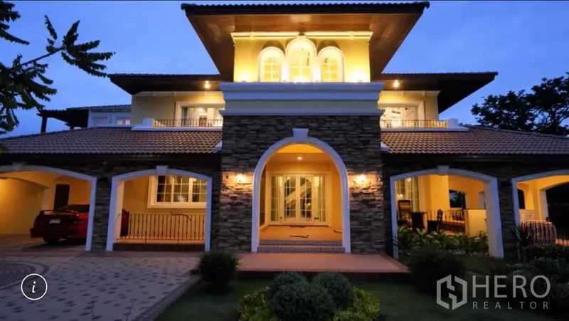 Detached House for rent or sale in Bang Phli, Samut Prakan - Evening exterior of a grand detached house with stone arches, balconies, warm lighting, covered carports and a manicured garden.