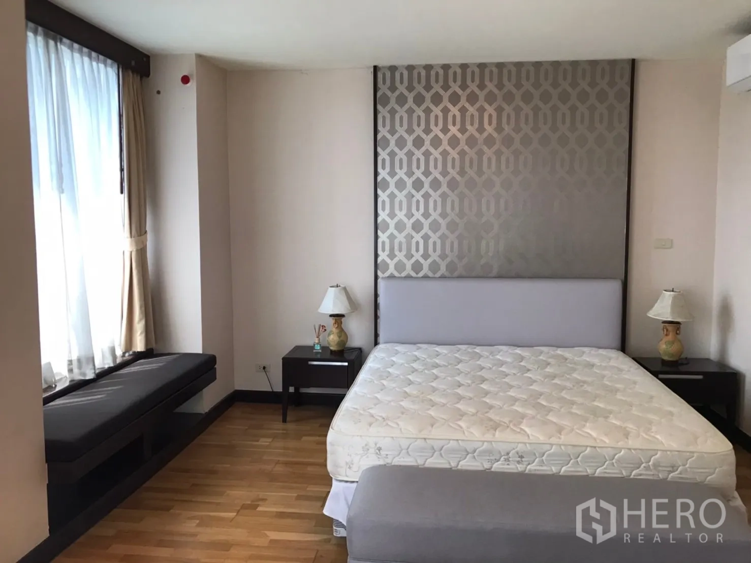 Condominium for rent or sale in Pathum Wan, Bangkok - Primary bedroom with upholstered headboard feature wall, bedside tables, bench seat by window and hardwood flooring.