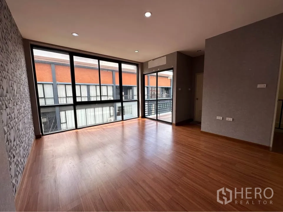 Townhouse for rent in Pak Kret, Nonthaburi - Spacious living area with wood flooring, floor-to-ceiling windows, and balcony access.