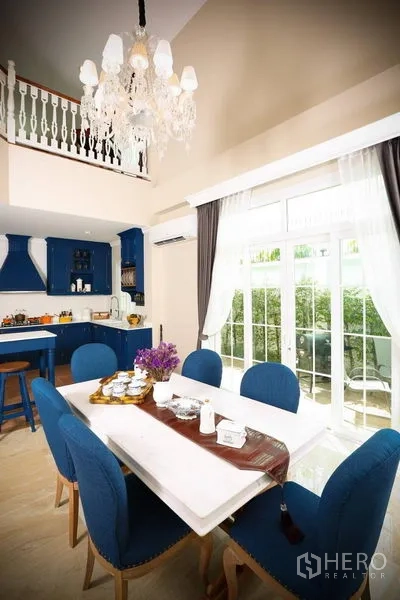 Detached House for rent in Bang Na, Bangkok - Dining zone under crystal chandelier with double-height ceiling, glass doors and adjoining blue kitchen.