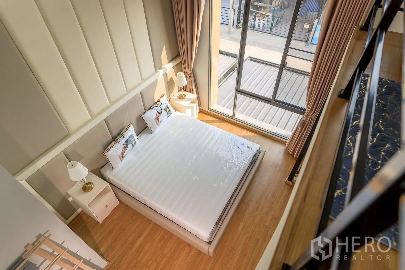 Townhouse for rent or sale in Bang Phli, Samut Prakan - Double-height master bedroom with king bed, large sliding glass windows and wood floors, viewed from the mezzanine.