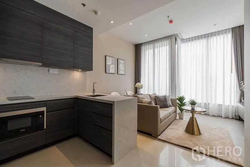 Condominium for rent in Watthana, Bangkok - Modern open-plan kitchen with dark cabinetry flowing into a bright living area with floor-to-ceiling windows.