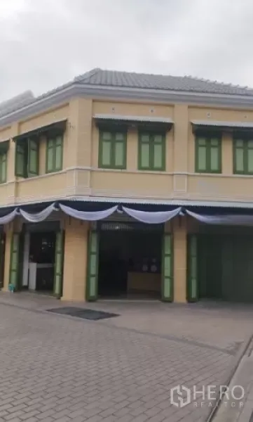 零售空间 出租在Bangkok - 两层转角骑楼式店面，米色外立面与绿色百叶窗，门面醒目。