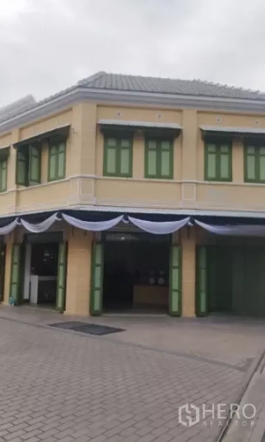 零售空间 出租在Bangkok - 两层转角骑楼式店面，米色外立面与绿色百叶窗，门面醒目。