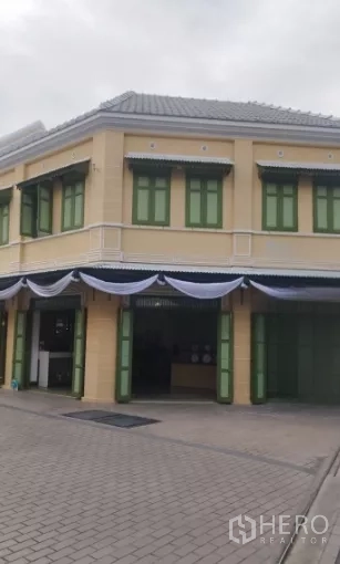 零售空间 出租在Bangkok - 两层转角骑楼式店面，米色外立面与绿色百叶窗，门面醒目。