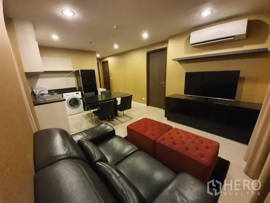 Condominium for rent in Sathon, Bangkok - Living area with black leather sofa, red ottomans, wall-mounted TV, dining set and kitchenette with washing machine.