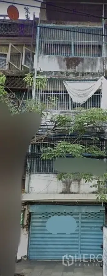 อาคารพาณิชย์ ให้เช่าในWatthana, Bangkok - อาคารพาณิชย์หลายชั้นด้านหน้า ประตูม้วนสีน้ำเงิน มีเหล็กดัด ระเบียง และผนังคอนกรีตเก่า พร้อมต้นไม้ริมถนนและสายไฟด้านหน้า